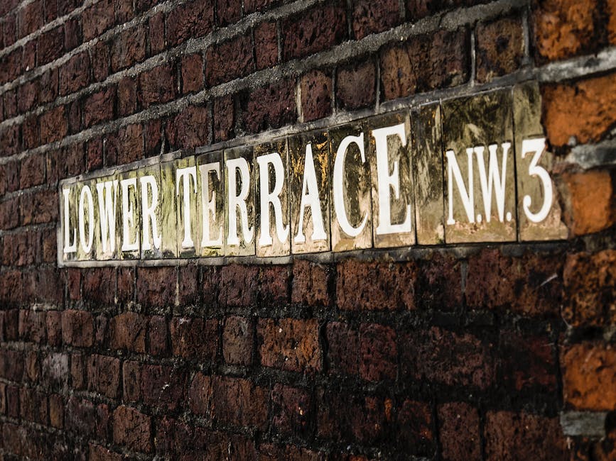 Close-up view of a rectangular street sign mounted on a red brick wall, displaying the text 'LOWER TERRACE NW3' in large white capital letters with a weathered appearance. The sign has a yellowish background with visible grime and moss, and the bricks surrounding it are dark red and brown with mortar joints in between. The sign is positioned slightly to the right within the frame, emphasizing its texture and aged condition. This scene depicts an urban residential area, and the image is relevant for house removals or home relocation services, illustrating a typical street view that might be encountered during furniture transport or packing and moving processes in Belsize Village, NW3, as provided by Man and Van Belsize Park.